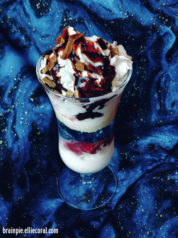 A tall glass with a knickerbocker parfait custard, which contains whipped cream, Jell-O, toasted nuts, fruit, vanilla ice cream, and chocolate syrup.