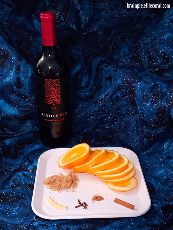 A bottle of red wine sits behind a white square plate filled with orange slices, a pile of brown sugar, a sliver of ginger, a cinnamon stick, cloves, nutmeg, all on a fabric decorated with blue and black starry galaxies.