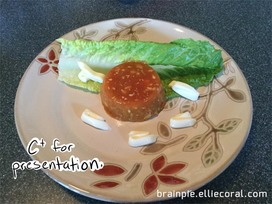 A small tomato aspic mold sits in the center of a china plate, surrounded with a single leaf of lettuce and some dabs of mayonnaise. It looks rather pathetic.