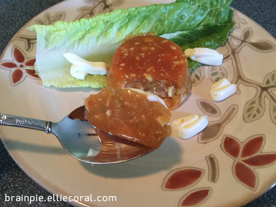 A spoon wields a portion of the tomato aspic, hovering above the plate.