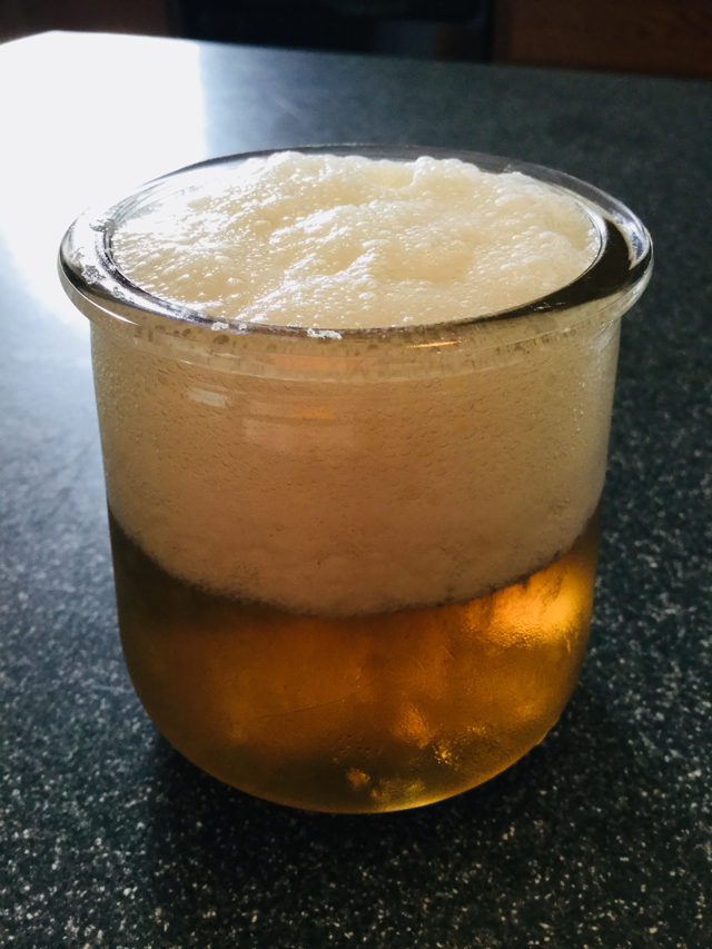 A small repurposed yogurt glass is filled with amber liquid with generous fizzy foam on top.