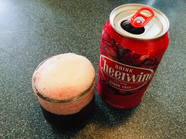 A small repurposed yogurt glass filled with dark cola and topped with pinkish fizzy foam. To the right is a Cheerwine can.