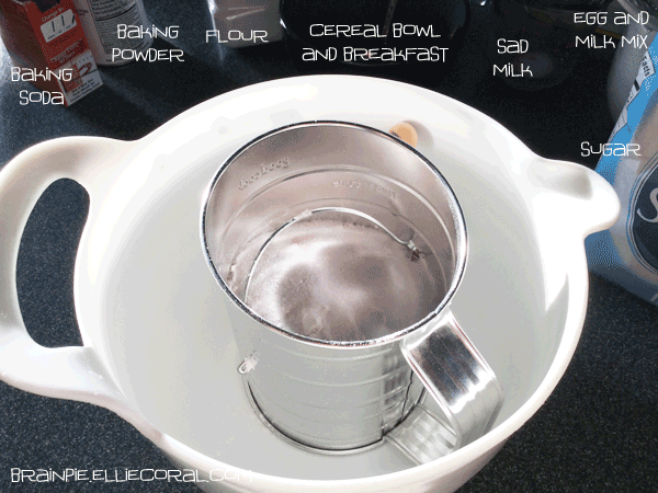 Another mixing bowl with a flour sifter. Off to the side are baking soda, baking powder, flour, a cereal bowl with breakfast, sad milk left alone in the measuring cup, egg and milk mix, and sugar.