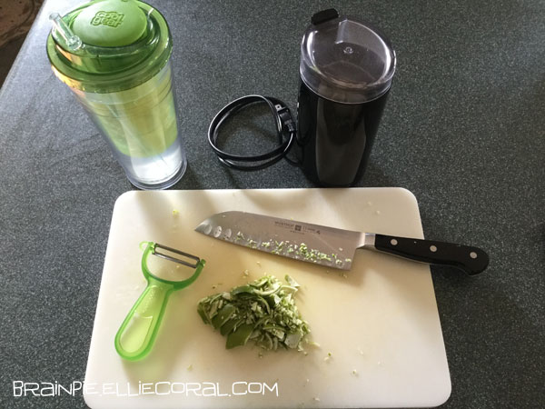 A tumbler with a strainer now houses the limes, which infuse the water with lime juice. A coffee grinder stands to the side. On the cutting board, there is a menacing looking knife, a carrot peeler, and some lime peels.