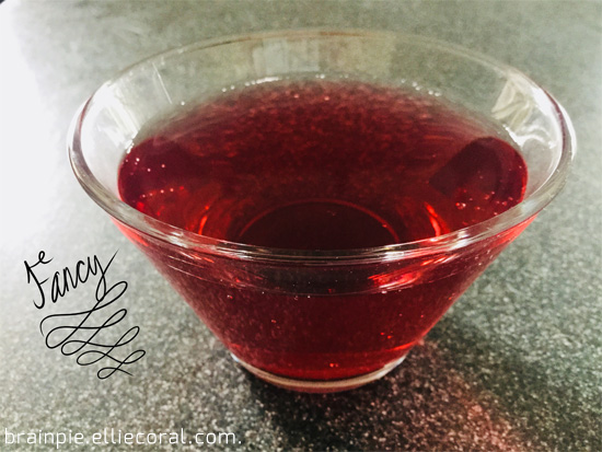 A triangular glass with a flat bottom holes clear red liquid. Some writing done in fancy script floats to the left and reads, 'Fancy'.