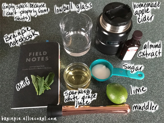 An assembly of tools: Brainpie notebook, mint, lowball glass, homemade apple cider, almond extract, sugar, lime, muddler, sparkling white grape juice.