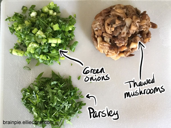 A cutting board with small piles of chopped green onions, thawed mushroom pieces, and parsley.