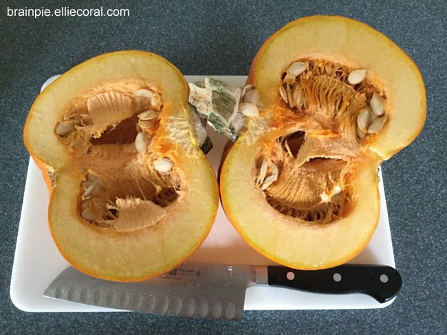 A pie pumpkin has been sliced in half, showing thick, light orange meat and stringy guts speckled with a few seeds. In the lower part of the image, a dangerous-looking carving knife sits on the cutting board.