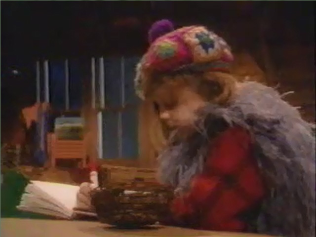 Bonnie sits at a desk, wearing a hat made from crocheted granny squares and a feather boa, as she writes in a journal.