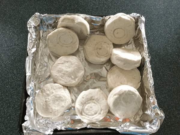 A square pan lined with aluminum foil with cold, uncooked biscuits thrown around carelessly.