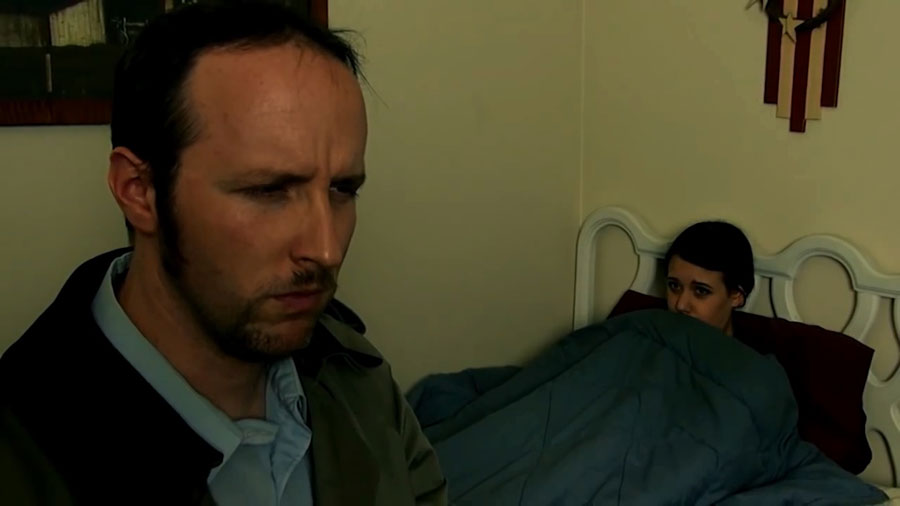 Doug Walker's character sits solemnly on the bed while Rachel's character sits up in bed with the covers clenched to her face.