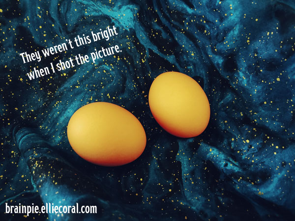 Two brown eggs sit on the blue galaxy fabric backdrop. They appear to have a yellow inner glow. The text reads, They weren't this bright when I shot the picture.