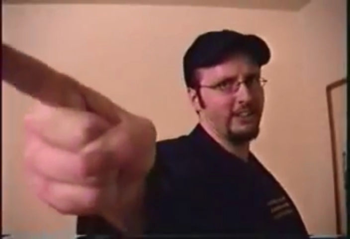 Doug Walker, dressed in a shirt, blazer, and baseball cap, points at the camera.